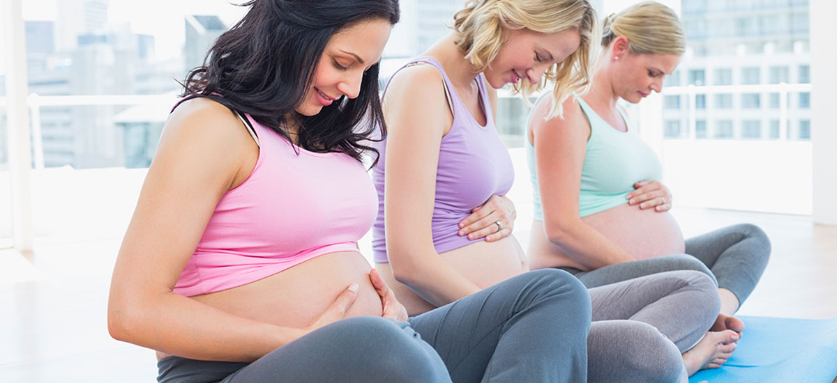 Three pregnant women sitting o yoga mats , holding their bellies and smiling
